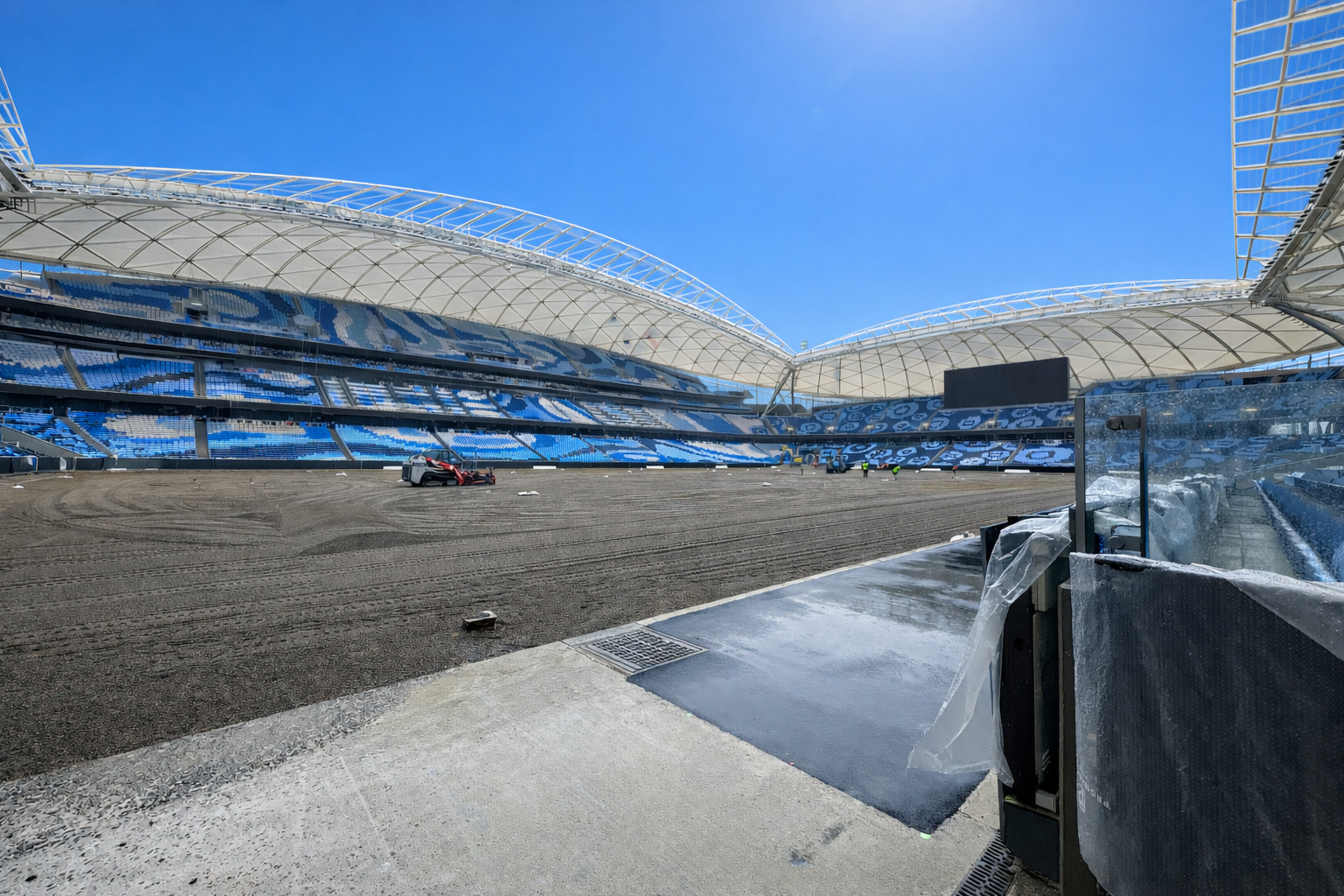 Empty stadium with blue and white seats, undergoing field renovation under a curved white roof on a clear sunny day.