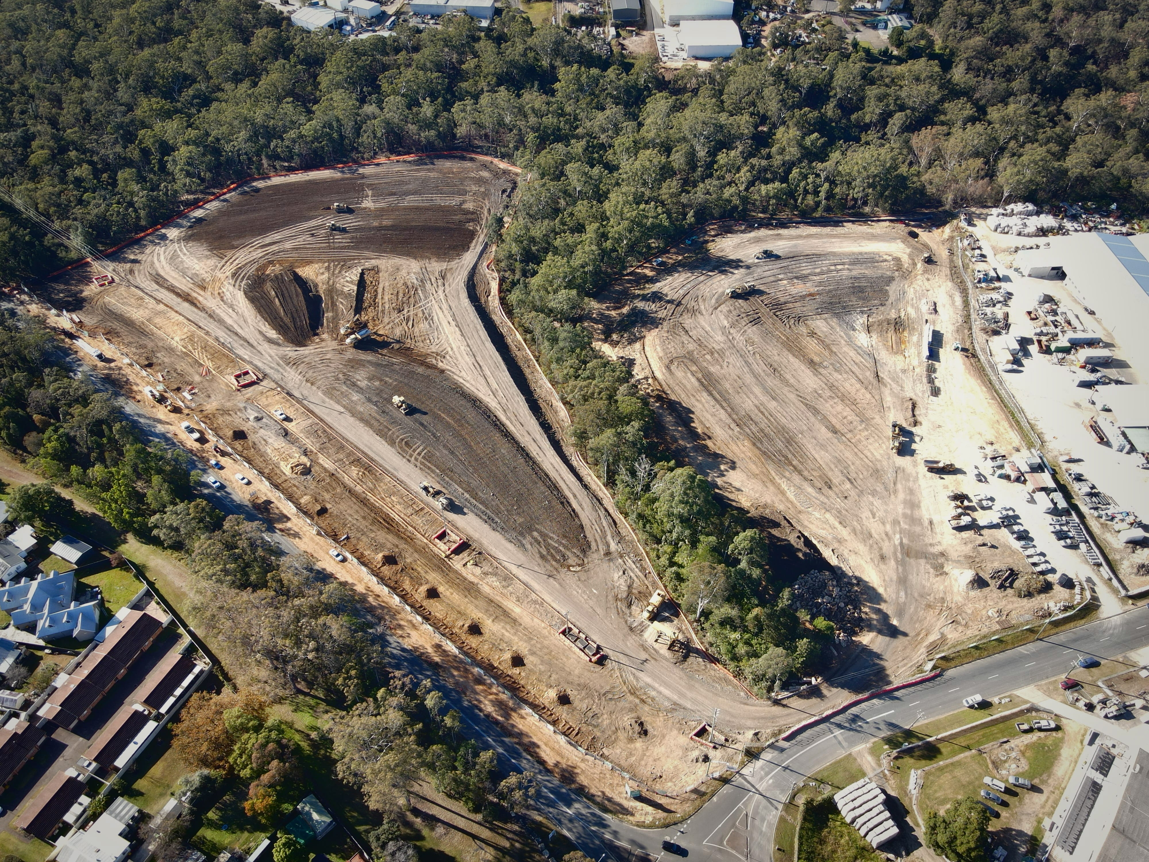 Aerial view of a large construction site clearing land with heavy machinery surrounded by forest and nearby buildings.