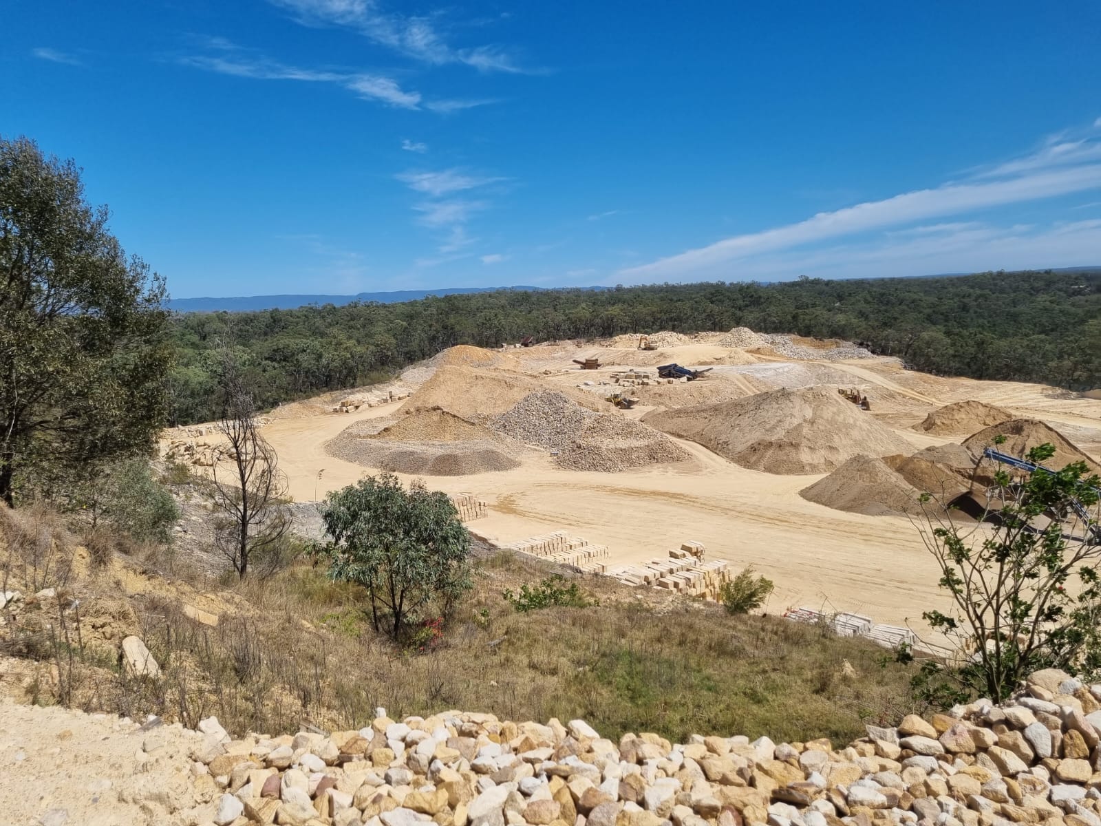 Construction site with piles of sand and gravel surrounded by trees under a blue sky.