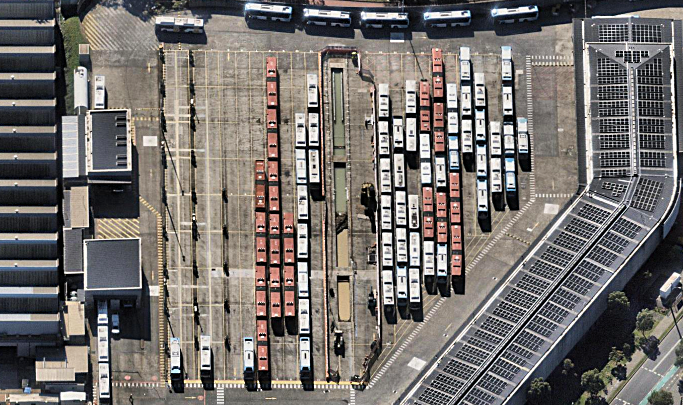 Aerial view of a bus depot with multiple parked buses arranged in rows between buildings with solar panels on the roof.