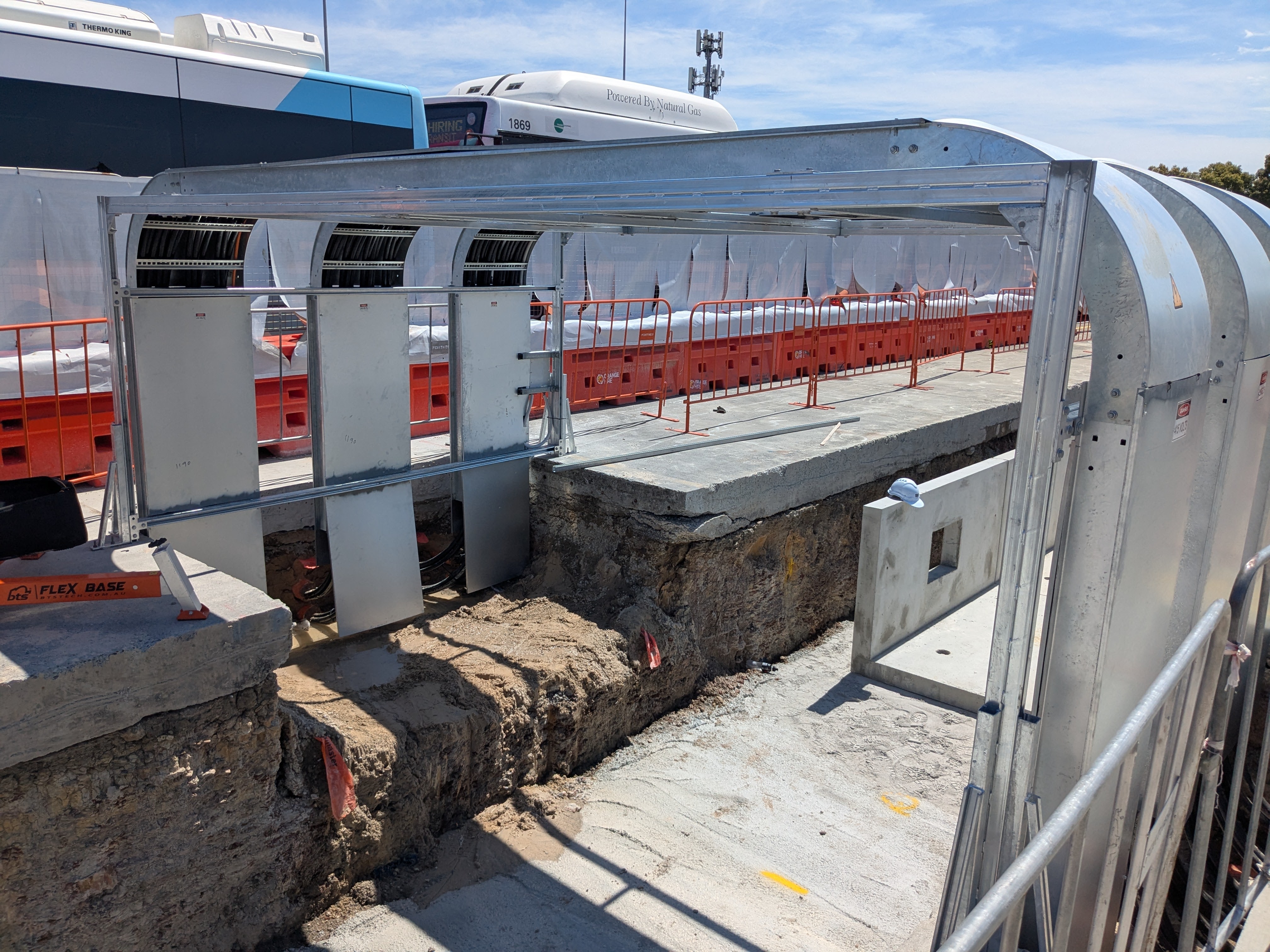 Construction site with exposed earth and concrete, metal barriers, and a covered metal walkway segment under a sunny sky with buses in the background.