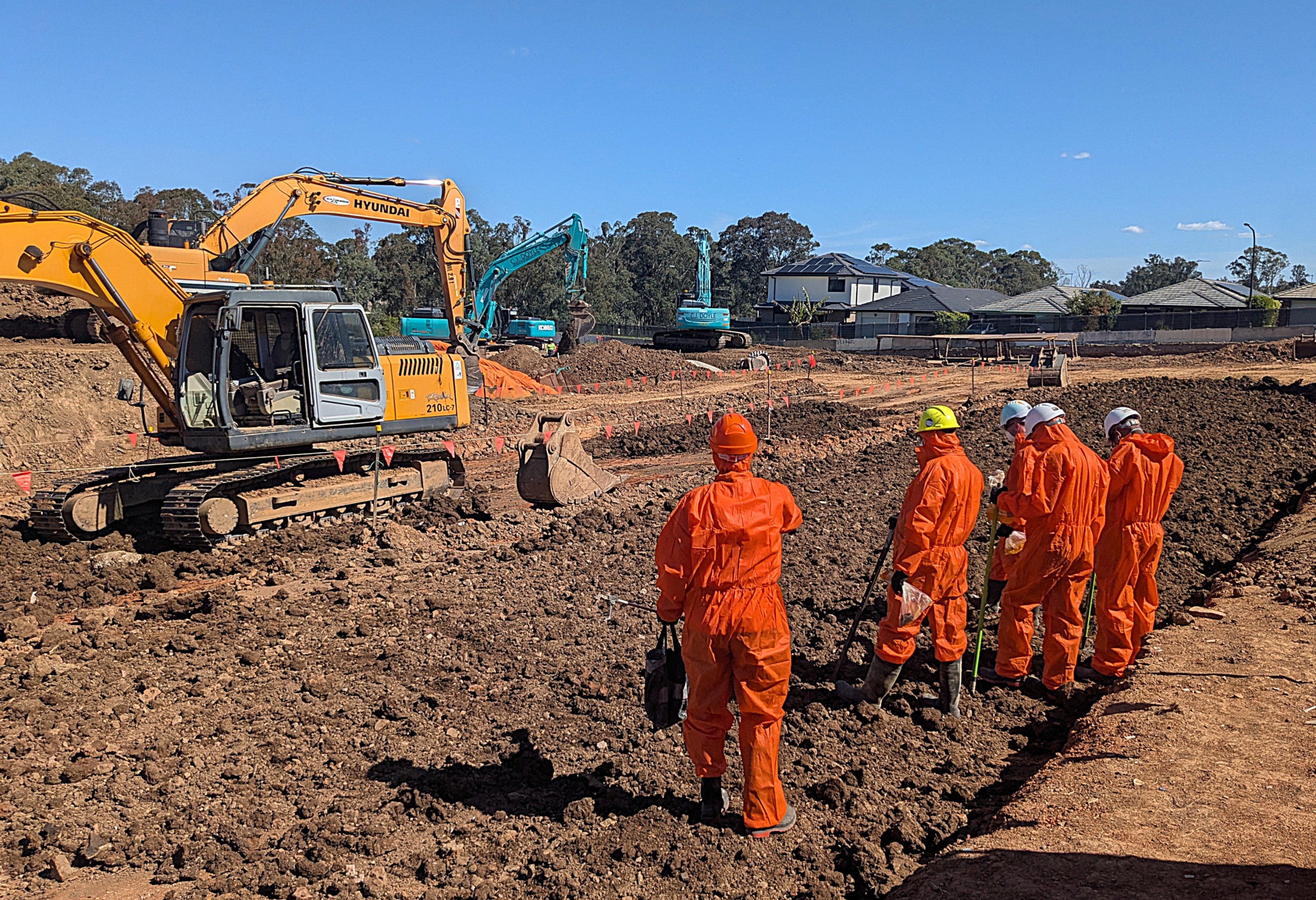 Five workers in orange protective suits and helmets standing on a muddy construction site near excavators.