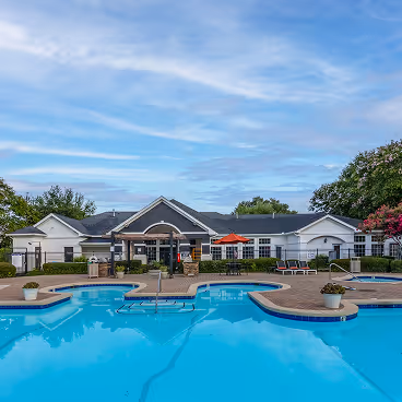 Outdoor swimming pool area with lounge chairs and a clubhouse building under a partly cloudy sky.