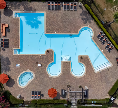 Aerial view of a uniquely shaped outdoor swimming pool with multiple lounging chairs and two red umbrellas around the pool area.