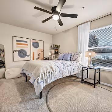 Cozy bedroom with a bed covered in a patterned light gray blanket, two framed abstract art pieces on the wall, a round beige rug, and a window with white blinds and curtains.