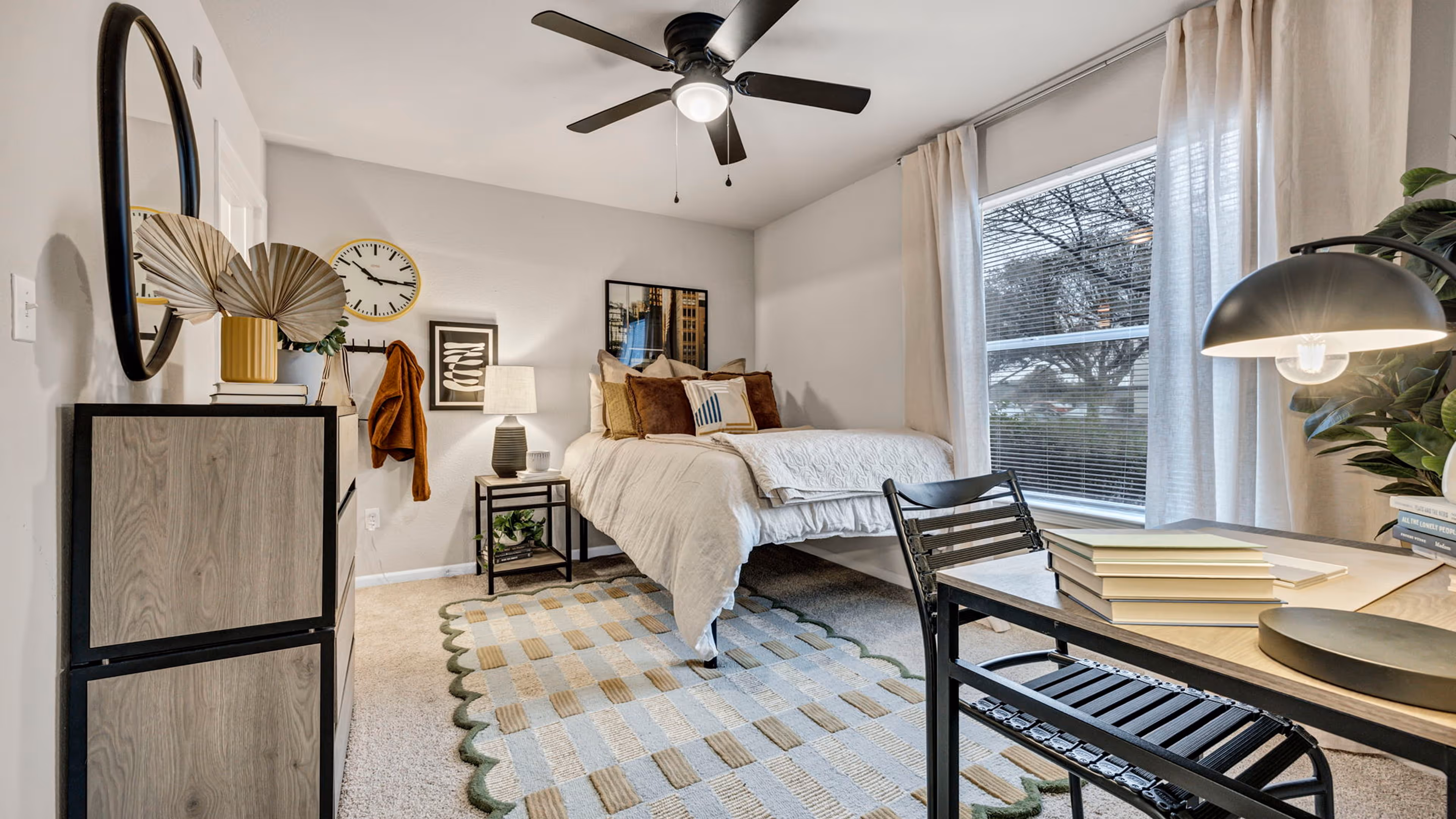 Modern bedroom with a neatly made bed, a patterned rug, a desk with a lamp and books, and a window with beige curtains.