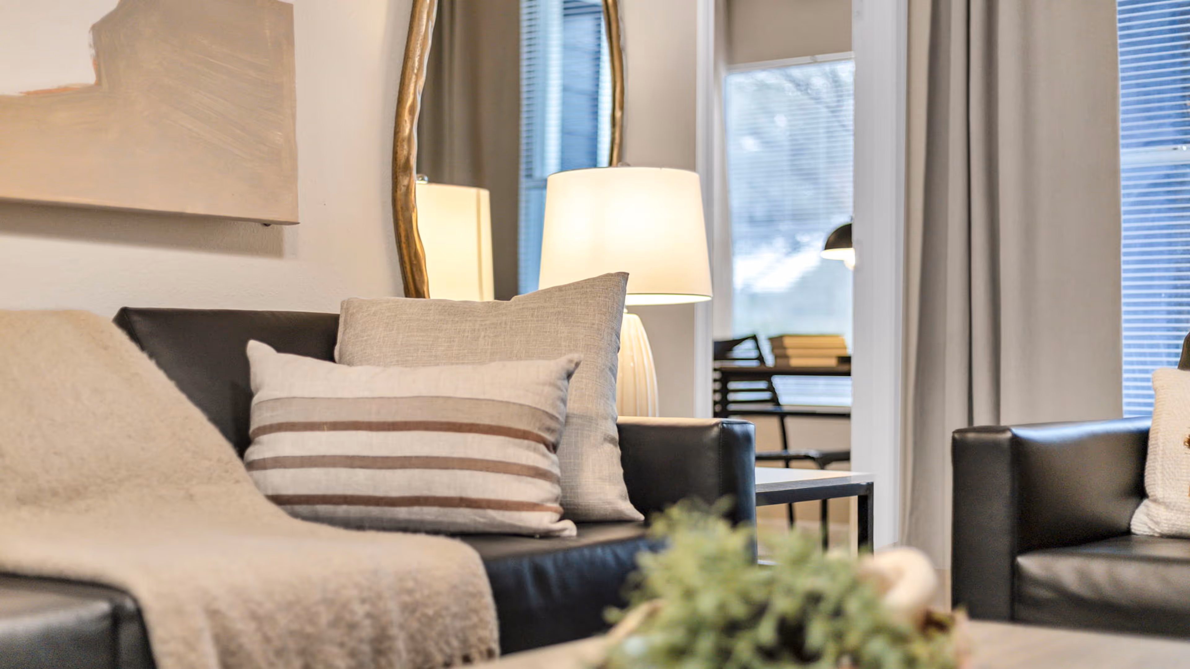 Cozy living room with black leather sofas, beige throw blanket, decorative pillows, a table lamp, and a gold-framed mirror.