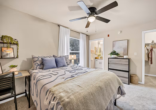 Cozy bedroom with a queen bed dressed in patterned bedding and blue pillows, a ceiling fan, a dresser, and an open doorway leading to a bathroom and closet.