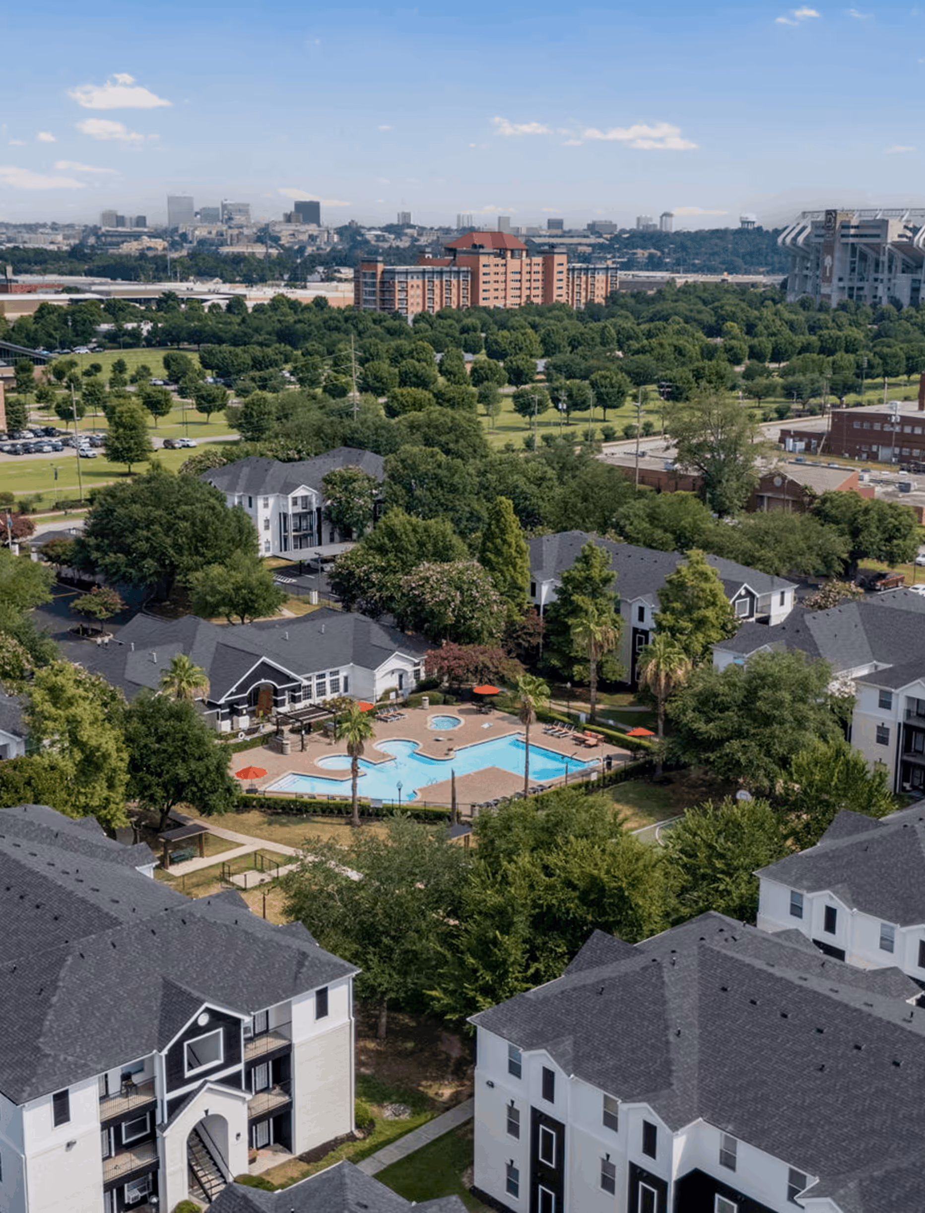 Aerial view of an apartment complex with a swimming pool, surrounded by trees and multiple buildings with dark roofs.