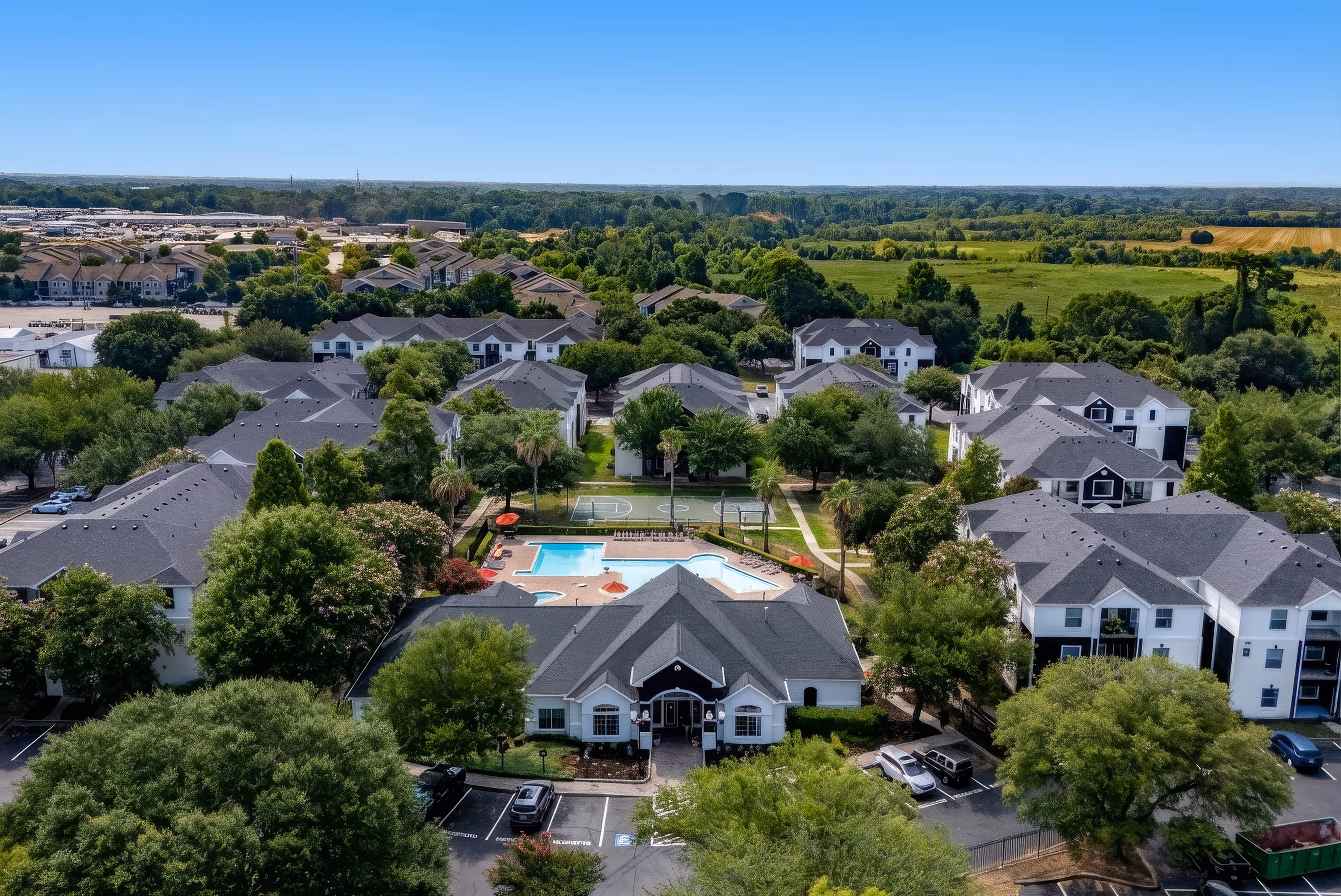 Aerial view of a residential apartment complex with multiple buildings, a central swimming pool, basketball court, surrounding trees, and parking lots.