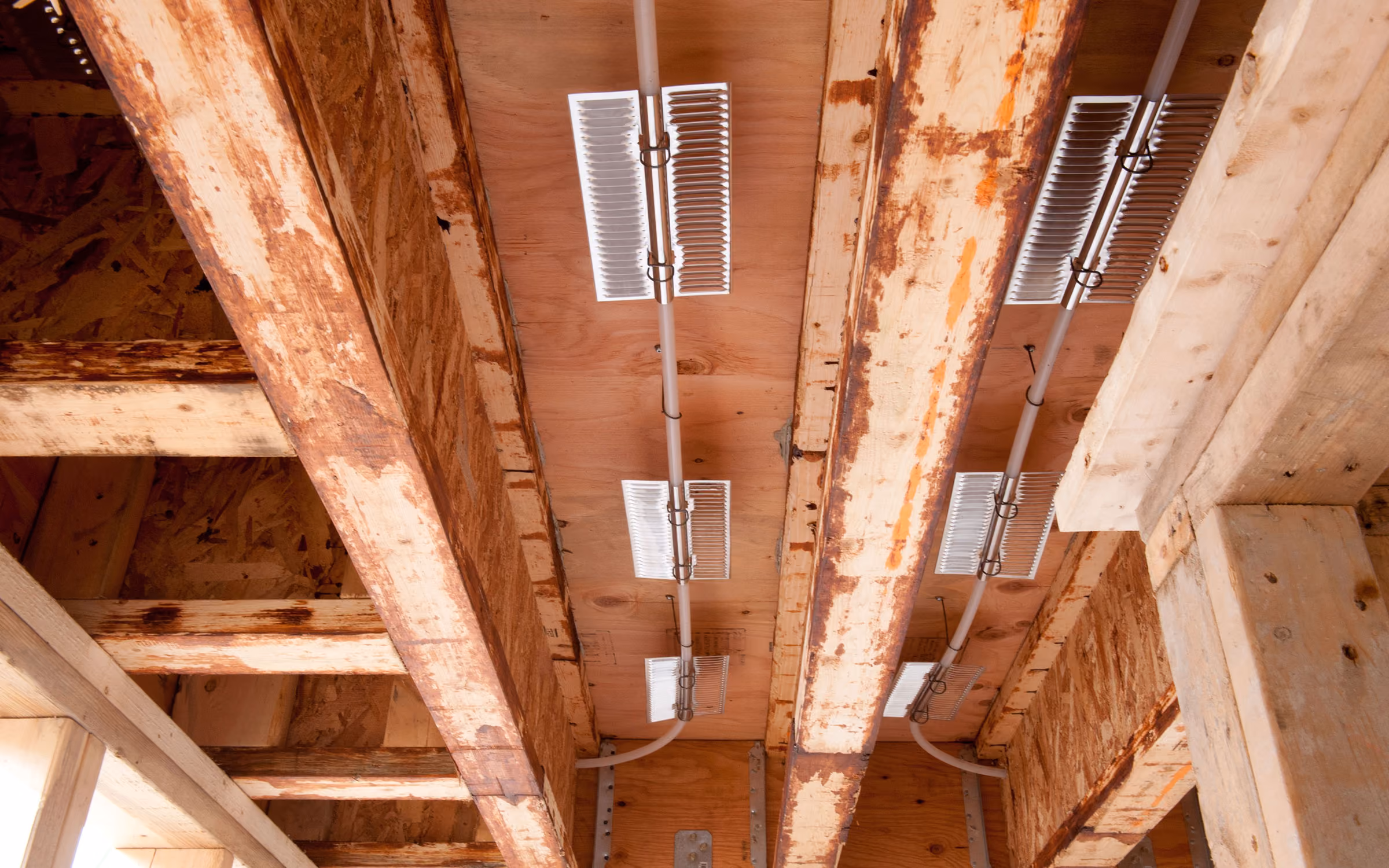 Exposed wooden ceiling beams with mounted heating panels and electrical wiring in a building under construction.
