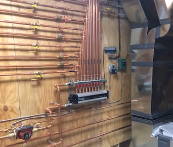 Copper piping system with valves and a control panel mounted on a wooden wall next to metal ductwork.
