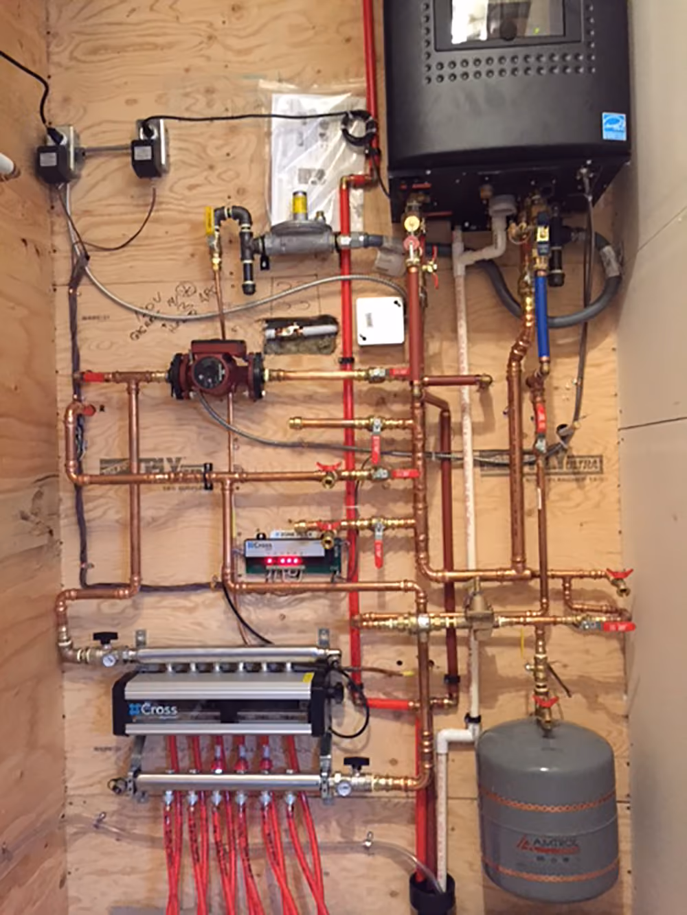 Complex heating system with copper pipes, valves, a pump, red tubing, and a grey expansion tank mounted on wood.