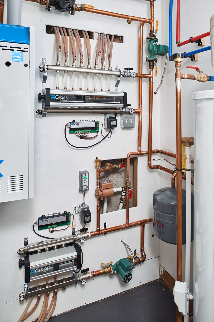 Indoor boiler system with copper pipes, Cross manifolds, pumps, and control panels mounted on a white wall.