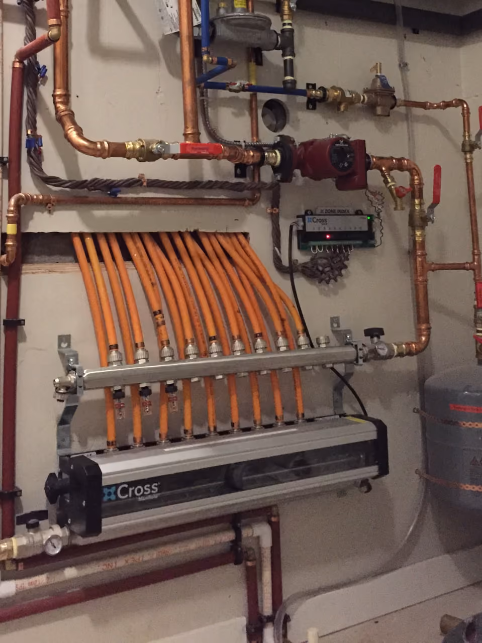 Wall-mounted plumbing system with copper pipes, orange flexible tubing, a Cross manifold, and control valves.