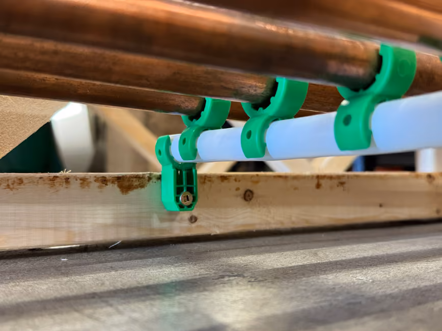 Green pipe clips securing white and copper pipes to a wooden frame against a concrete floor.