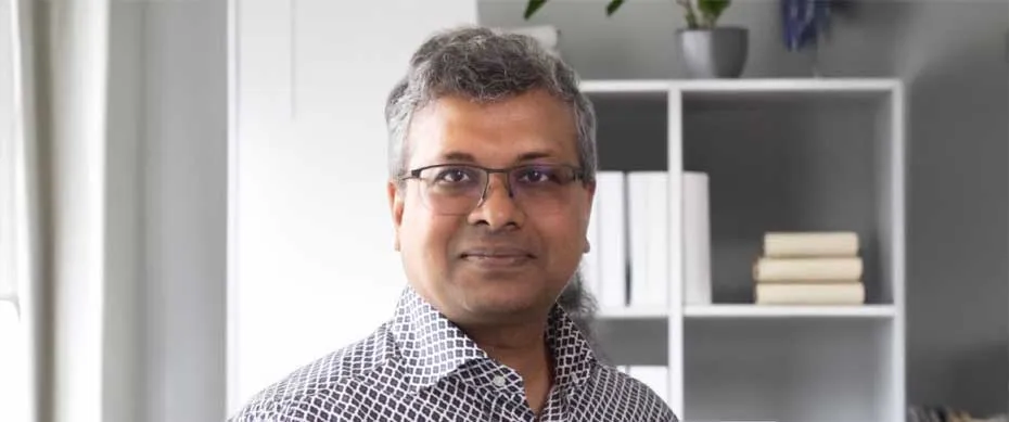 Smiling man with glasses, short gray hair, and patterned shirt in a modern room with a white bookshelf holding books and plants in the background.
