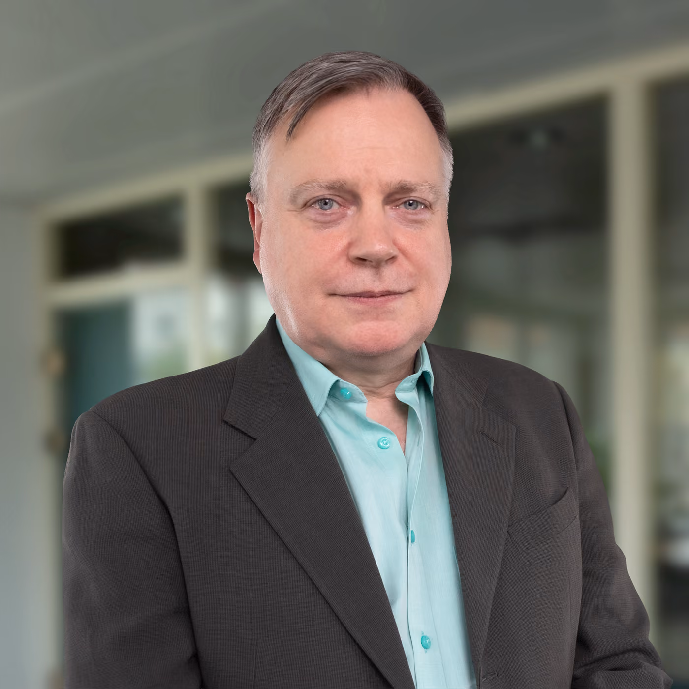 Middle-aged man with light skin and short grayish hair wearing a teal shirt and dark blazer standing indoors with a blurred background.