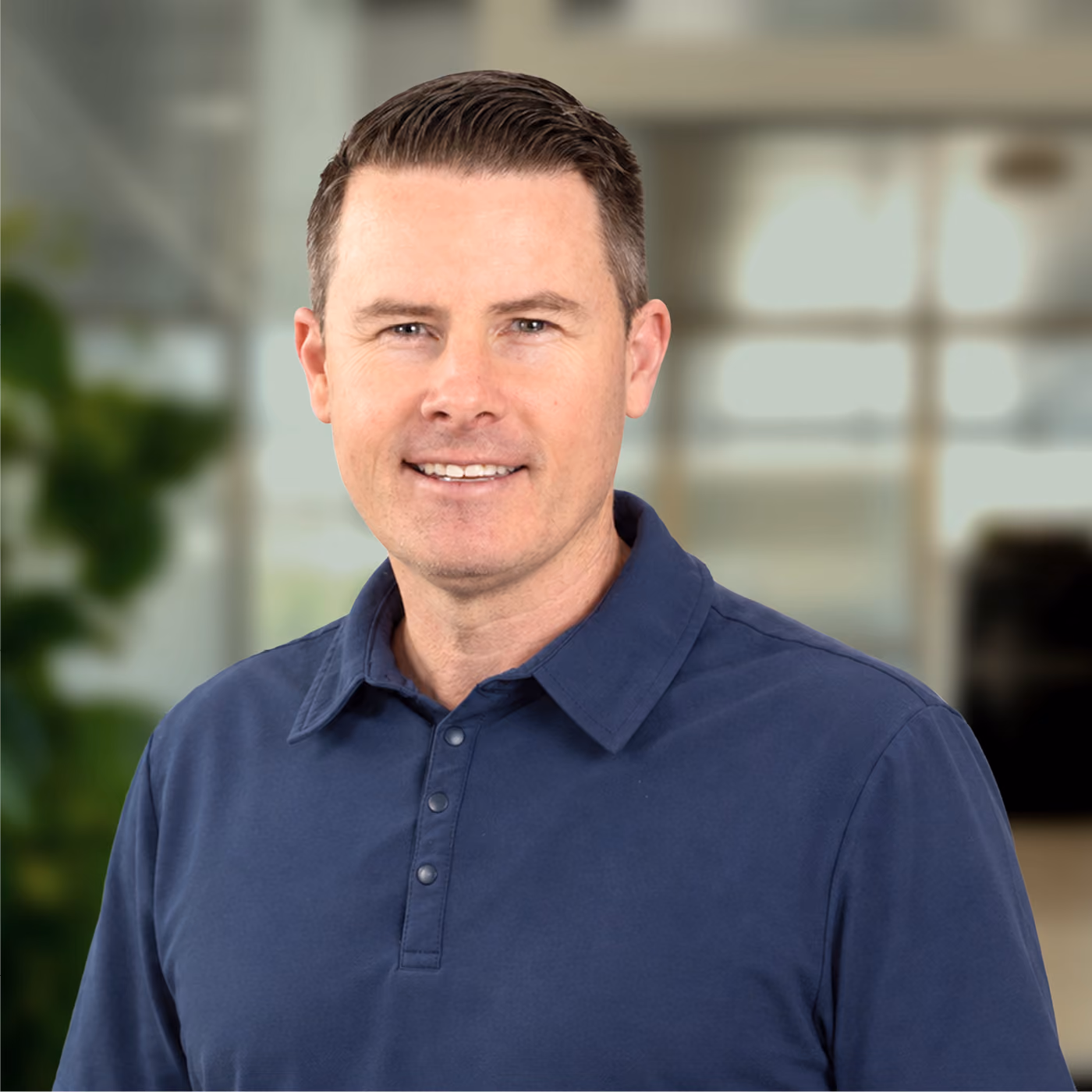 Smiling man with short dark hair wearing a navy blue collared shirt in a softly blurred indoor setting with greenery and light from windows.