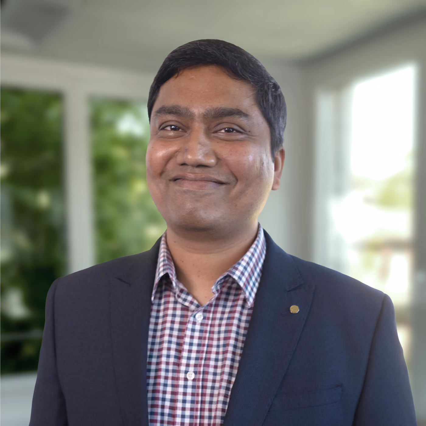Smiling man with short dark hair wearing a dark blazer and a checkered shirt standing indoors with blurred windows and greenery in the background.