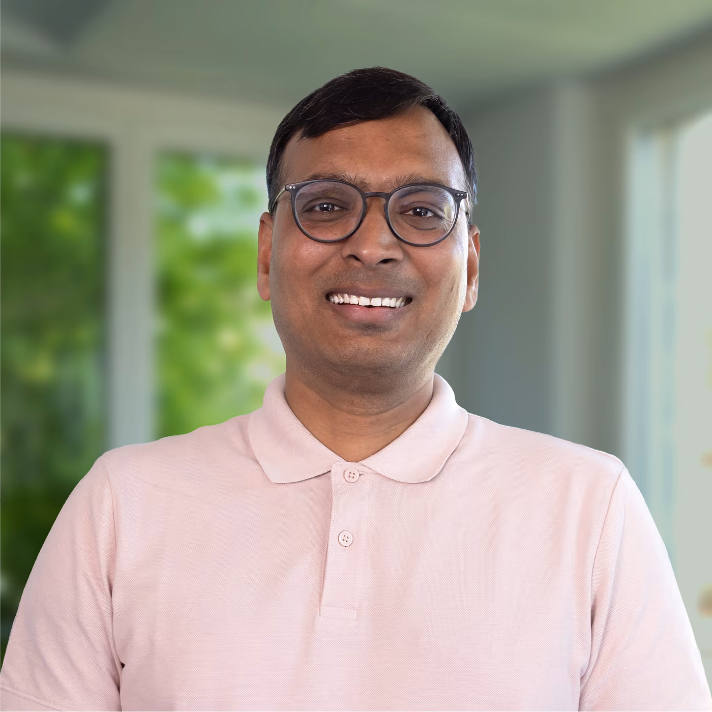 Smiling man wearing glasses and a light pink polo shirt standing indoors with blurred windows and green foliage in the background.