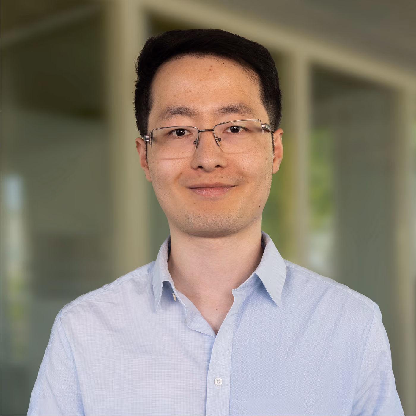 Smiling man with short black hair and glasses wearing a light blue button-up shirt, standing indoors with a blurred background.