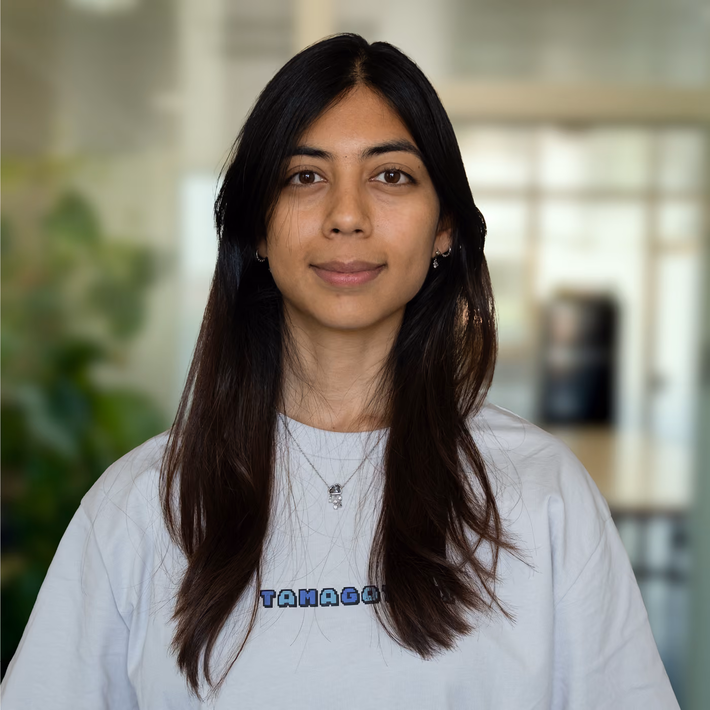 Young woman with long dark hair wearing a white T-shirt with the word 'TAMAGOTCHI' printed on it, standing indoors with a blurred background of windows and plants.