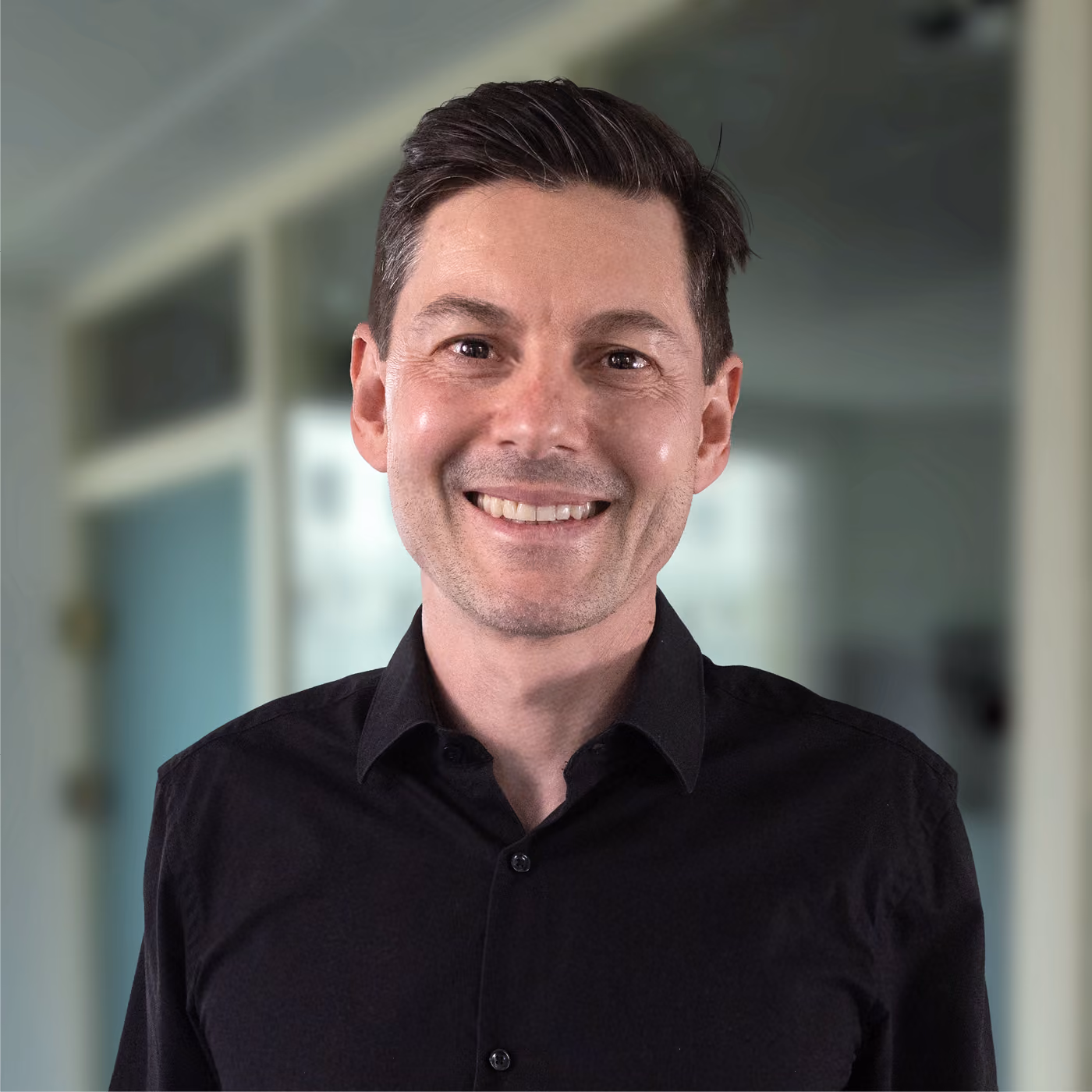 Smiling man with short dark hair and light skin wearing a black button-up shirt, standing in a softly blurred indoor setting.