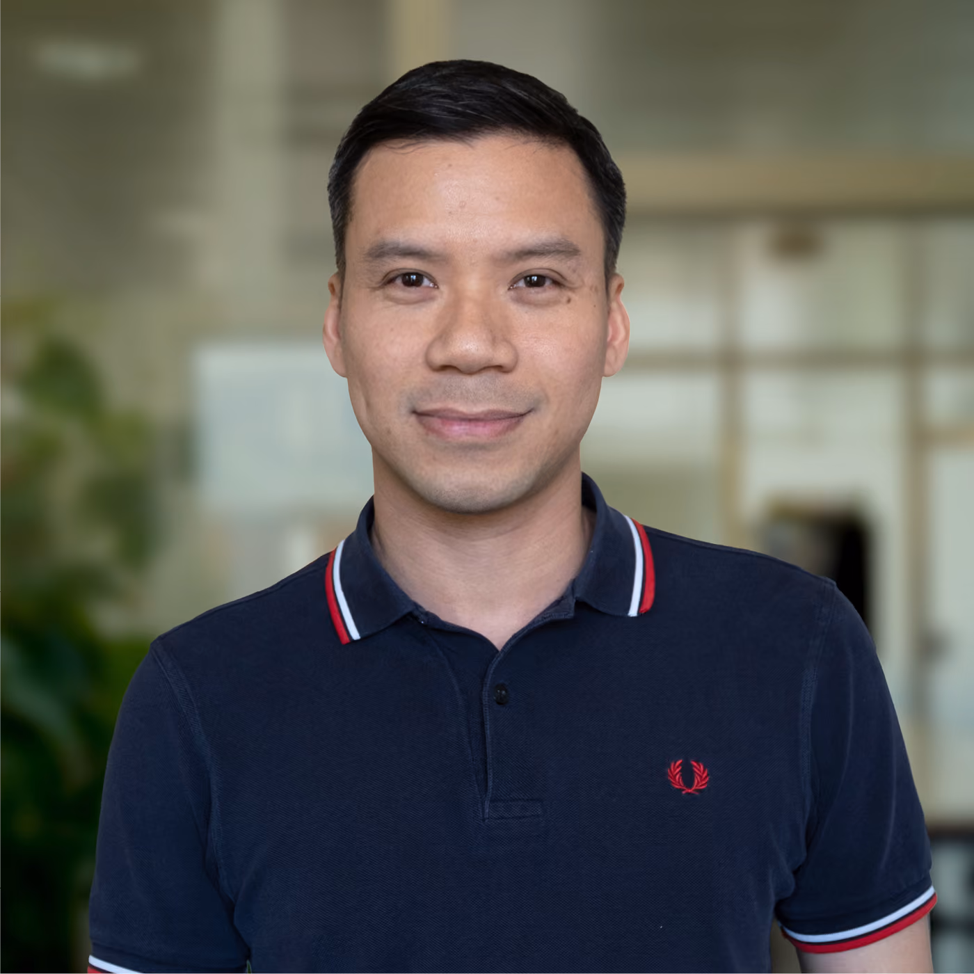 Portrait of a man with short black hair wearing a navy blue polo shirt with red and white trim on the collar and sleeves, standing indoors with a blurred background.