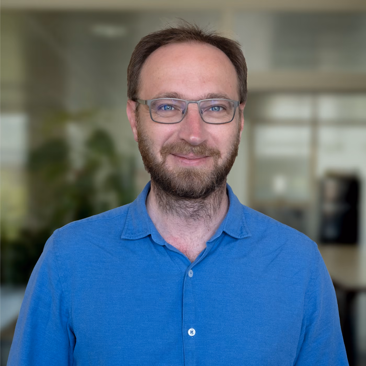 Smiling man with glasses, beard, and short brown hair wearing a blue button-up shirt, standing indoors with a blurred background.