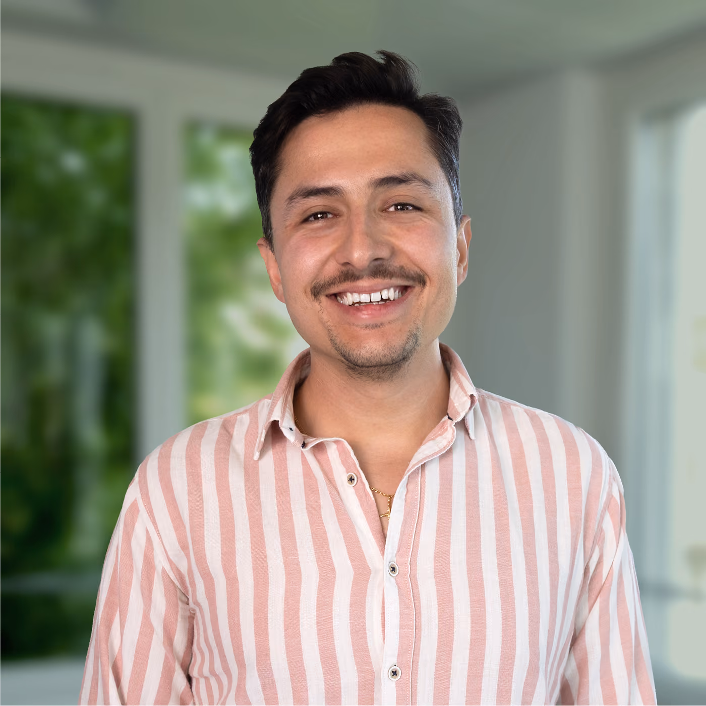 Smiling man with short dark hair and a mustache wearing a pink and white vertical striped shirt standing indoors with blurred green foliage and windows in the background.