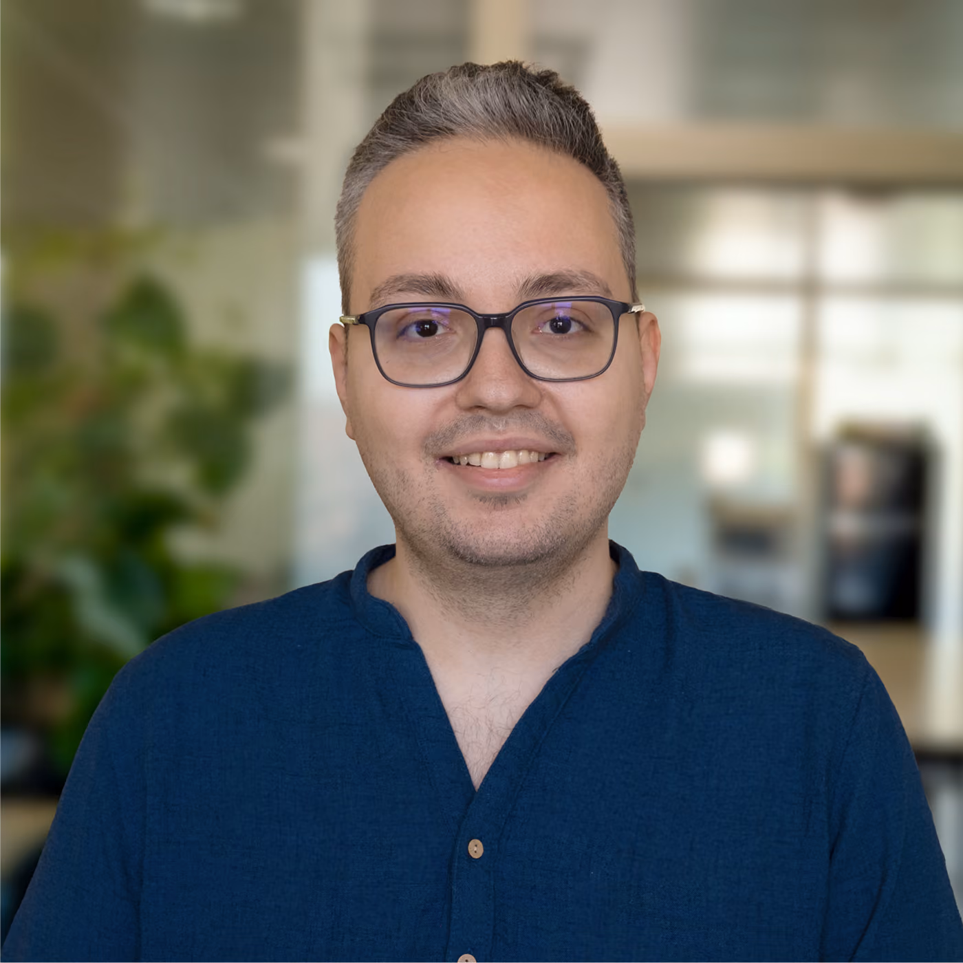 Smiling man with short salt-and-pepper hair and glasses wearing a blue shirt, standing indoors with blurred background including plants and office elements.
