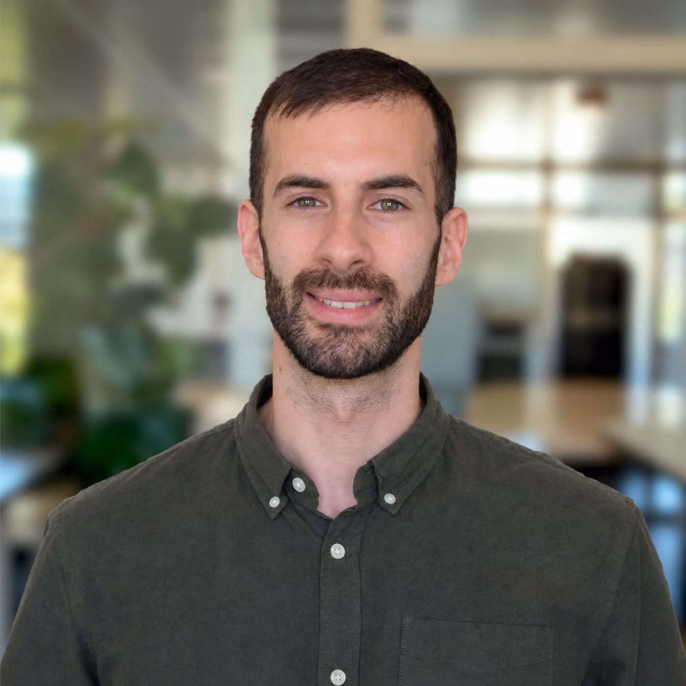 Smiling man with short dark hair and beard wearing a dark green button-up shirt, standing in a softly blurred office environment with plants and windows.