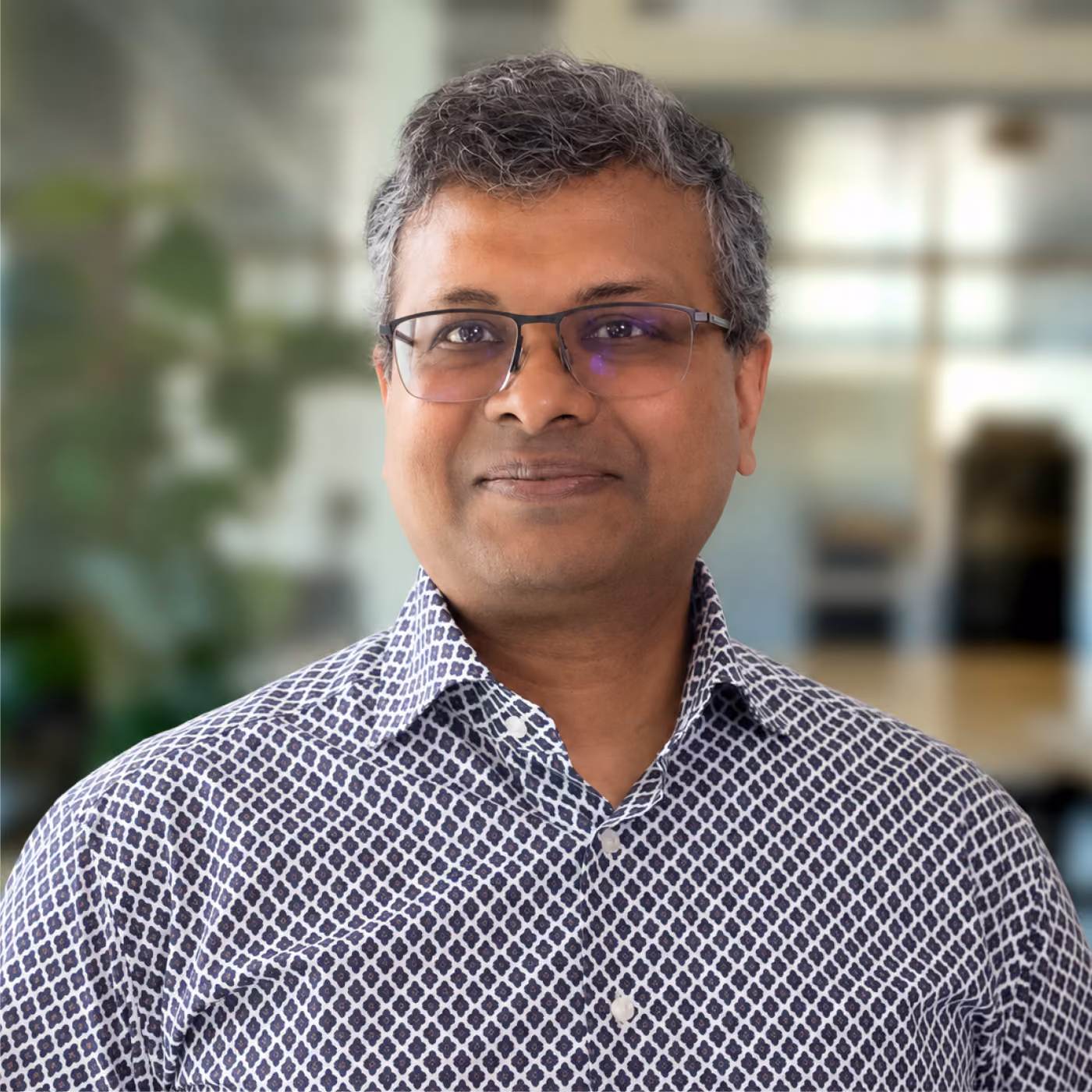 Smiling middle-aged man with gray hair wearing glasses and a patterned button-up shirt standing indoors with a blurred background.