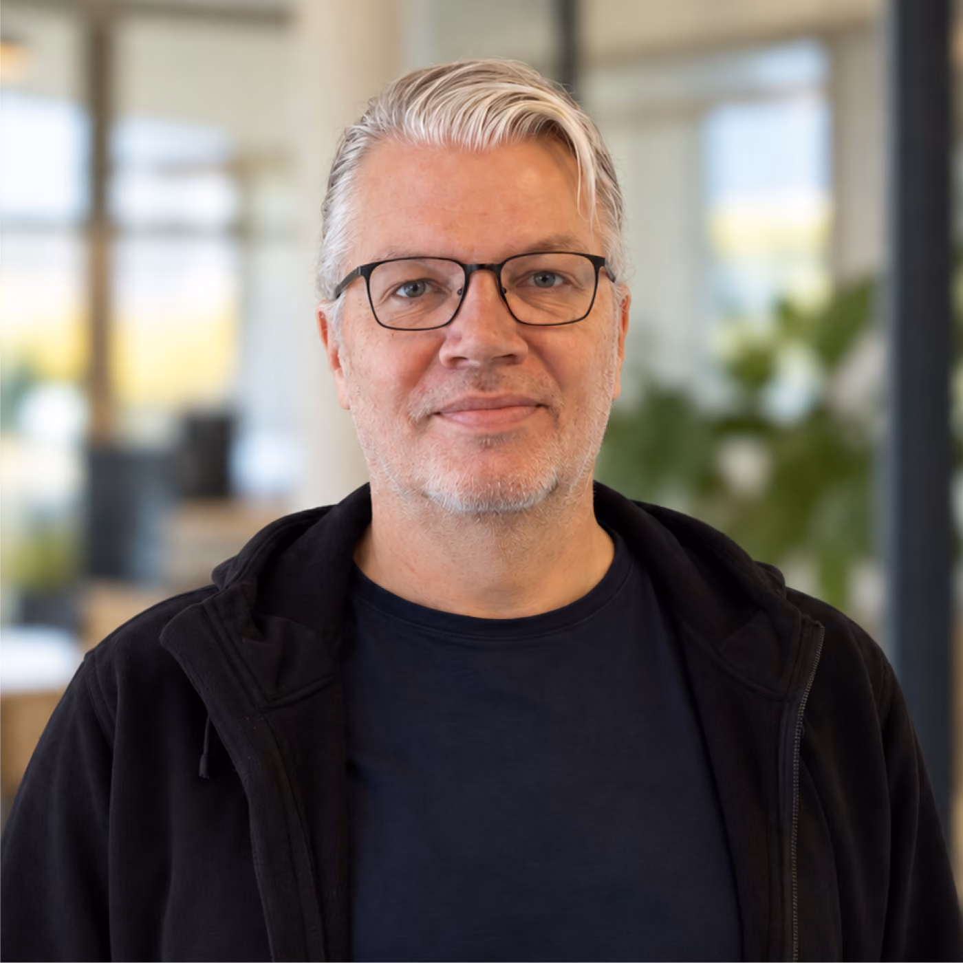 Middle-aged man with short white hair and glasses wearing a black jacket and navy blue shirt, standing indoors with a blurred office background.