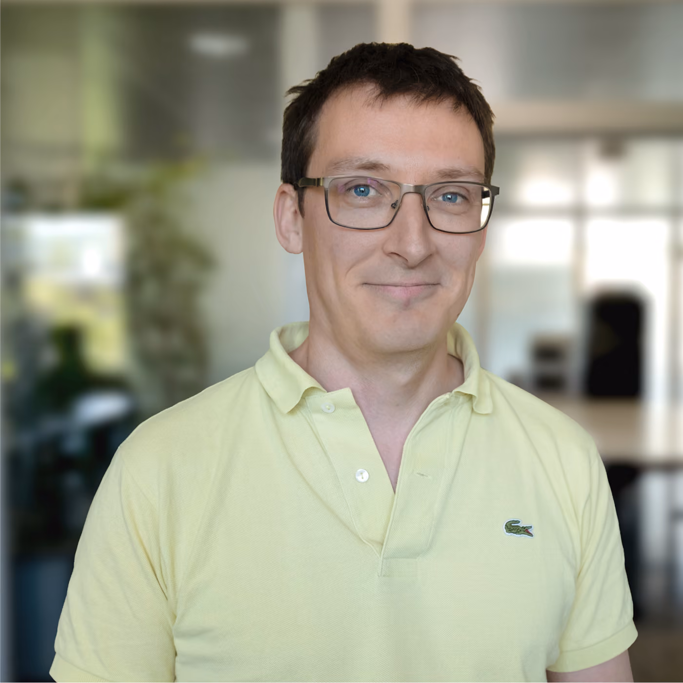 Smiling man with short brown hair and glasses wearing a light yellow polo shirt standing indoors with a blurred office background.