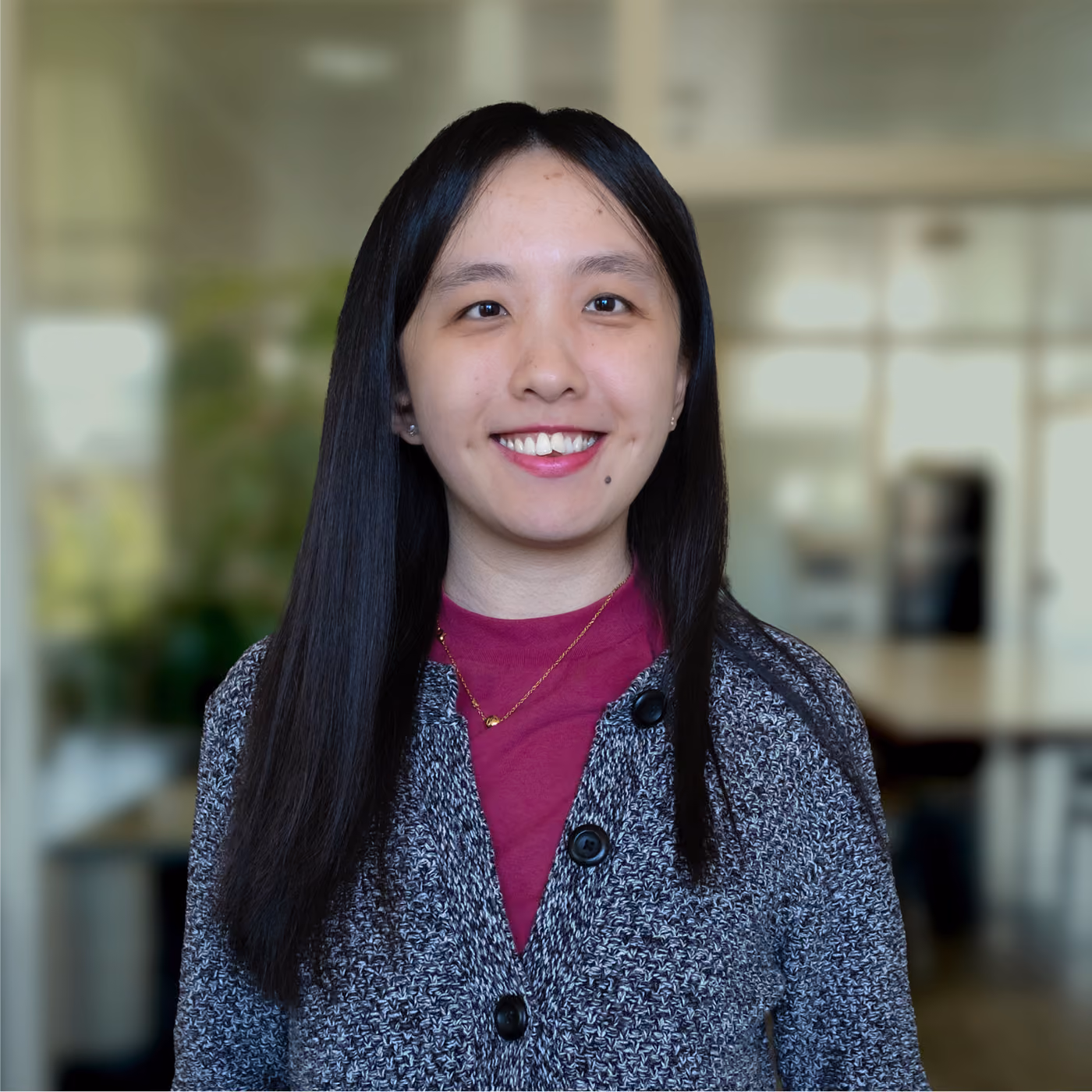 Smiling woman with long black hair wearing a gray knit cardigan over a maroon top and a gold necklace, standing in an office with blurred background.