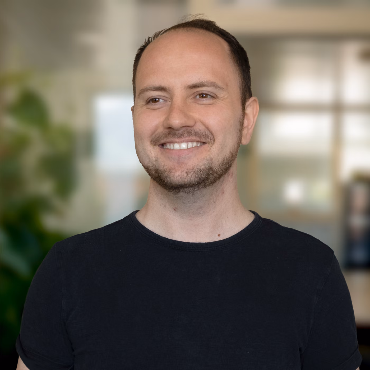 Smiling man with short dark hair and trimmed beard wearing a black t-shirt against a blurred indoor background with green plants and windows.