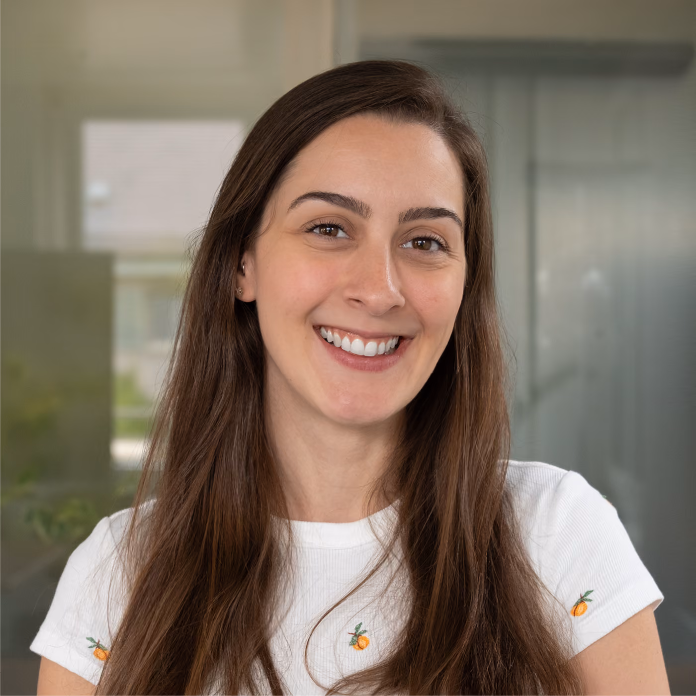 Smiling young woman with long brown hair wearing a white shirt with small embroidered oranges, standing indoors with blurred background.