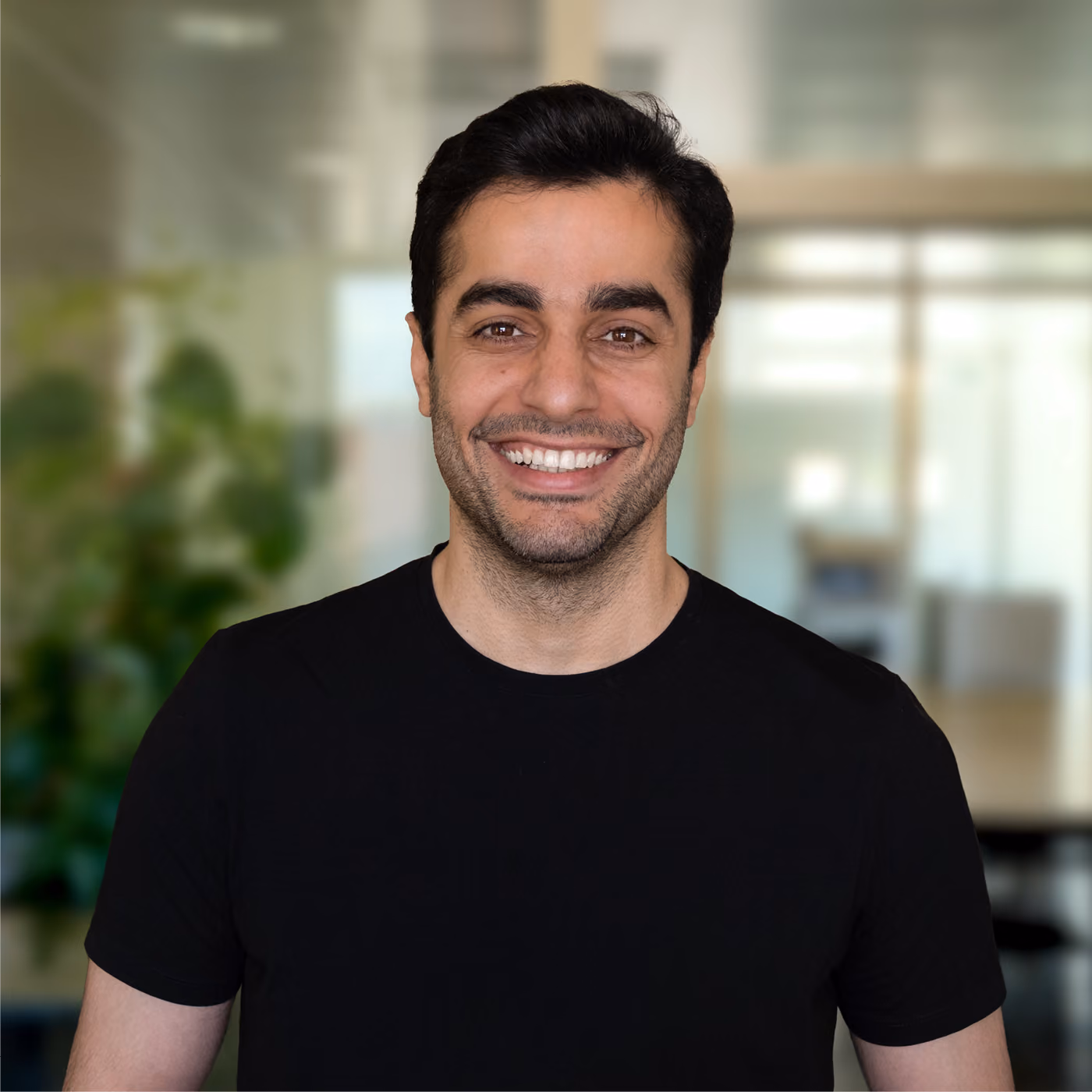 Smiling man with dark hair and beard wearing a black t-shirt, standing in a blurred office setting with plants and large windows.