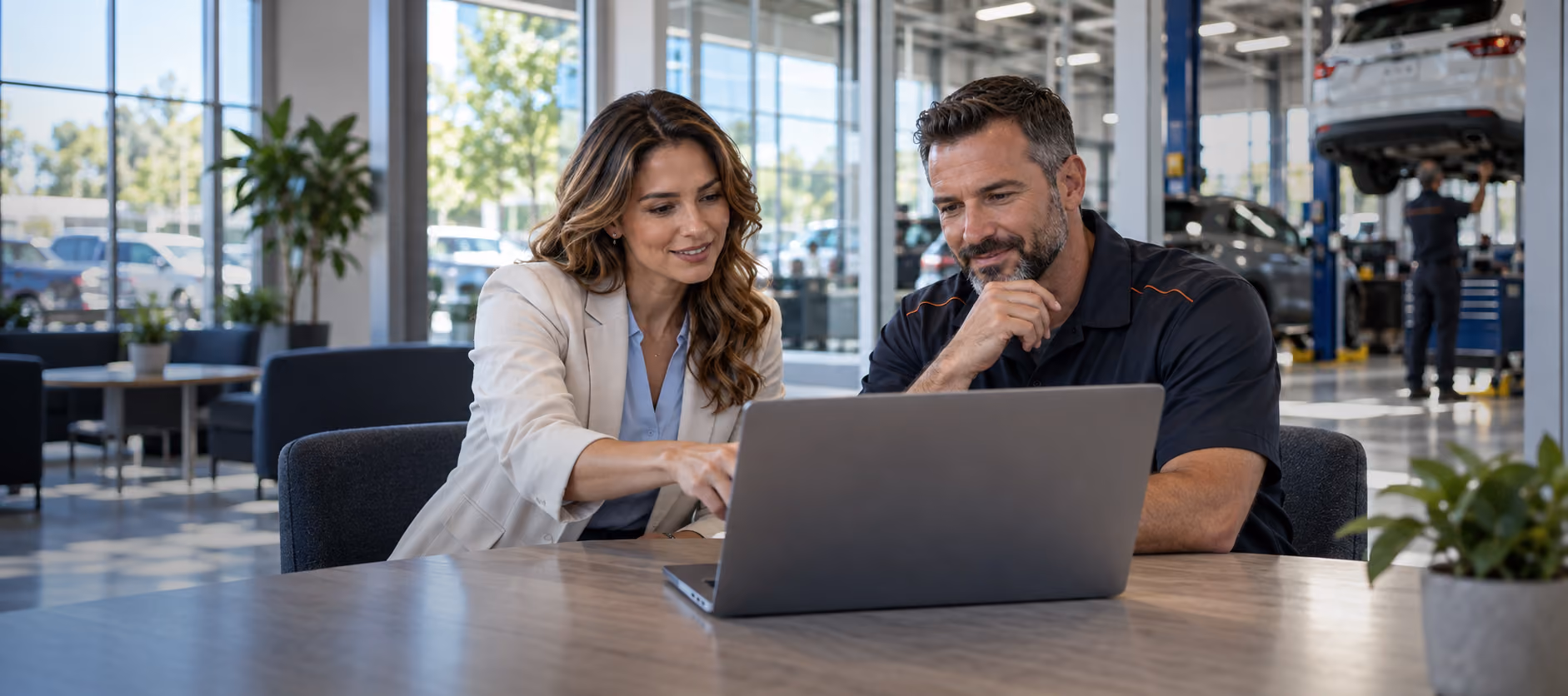 A man and a woman sitting at a table in a modern car service center, looking at a laptop as the woman points at the screen, while cars are being serviced in the background.