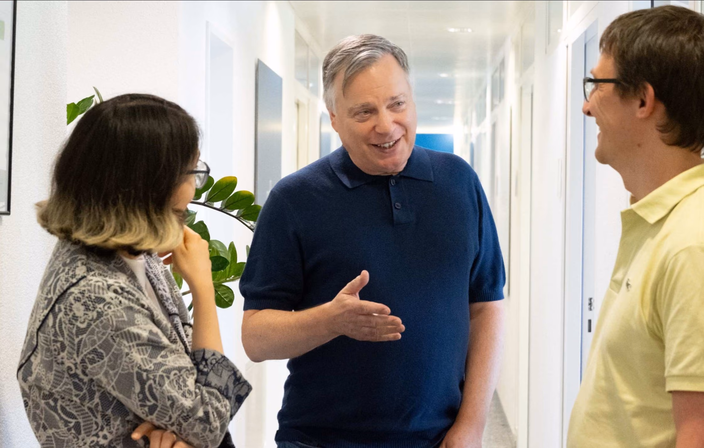 Three people having a friendly conversation in a bright hallway, with a man in a navy blue shirt gesturing while speaking, a woman with glasses and a patterned jacket listening thoughtfully, and a man in a pale yellow polo shirt smiling.