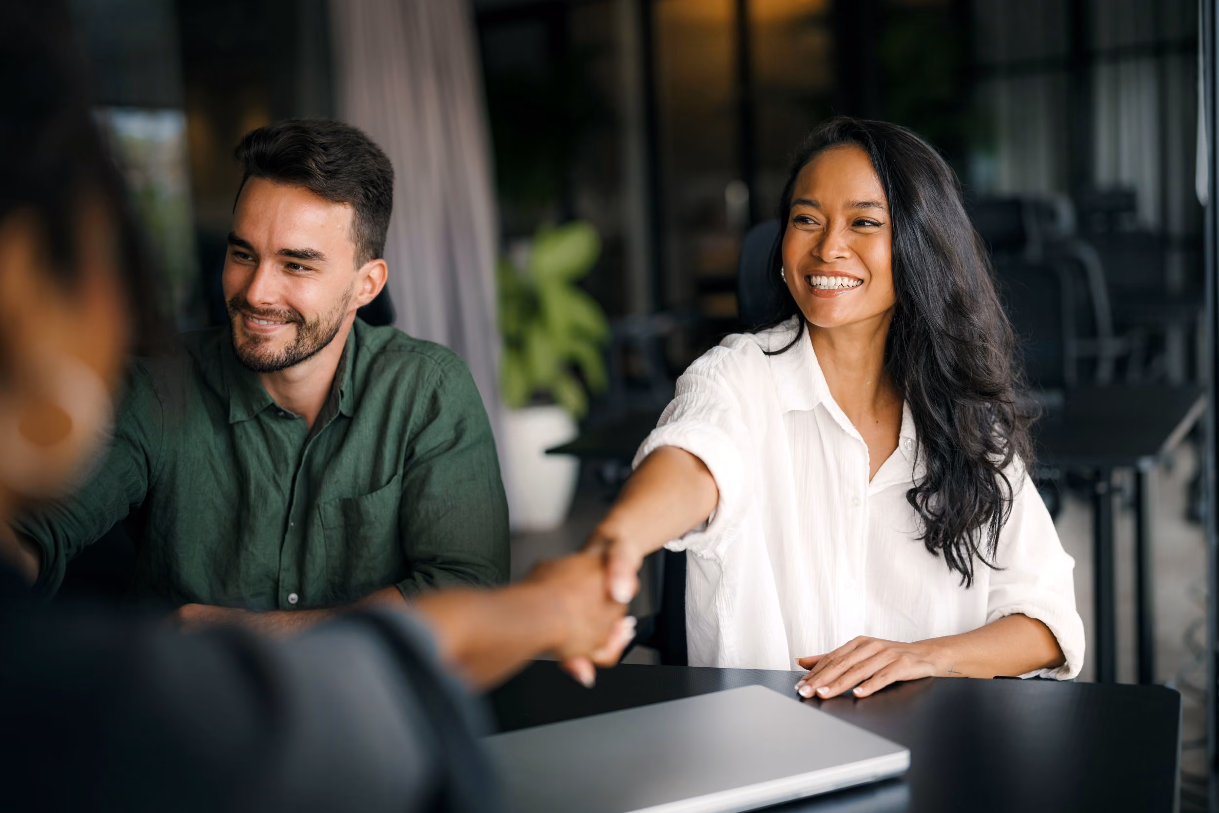 Smiling woman in a white shirt shaking hands with a person across the table, with a man in a green shirt watching and smiling, in a modern office setting.