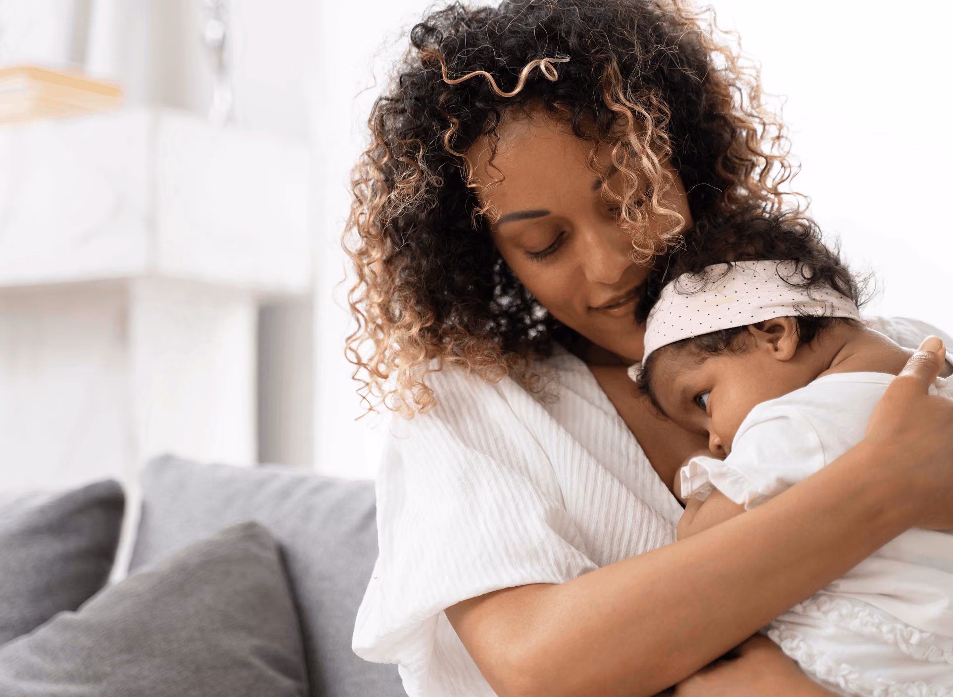 Mother gently holding and cuddling her baby wrapped in a white outfit and headband.
