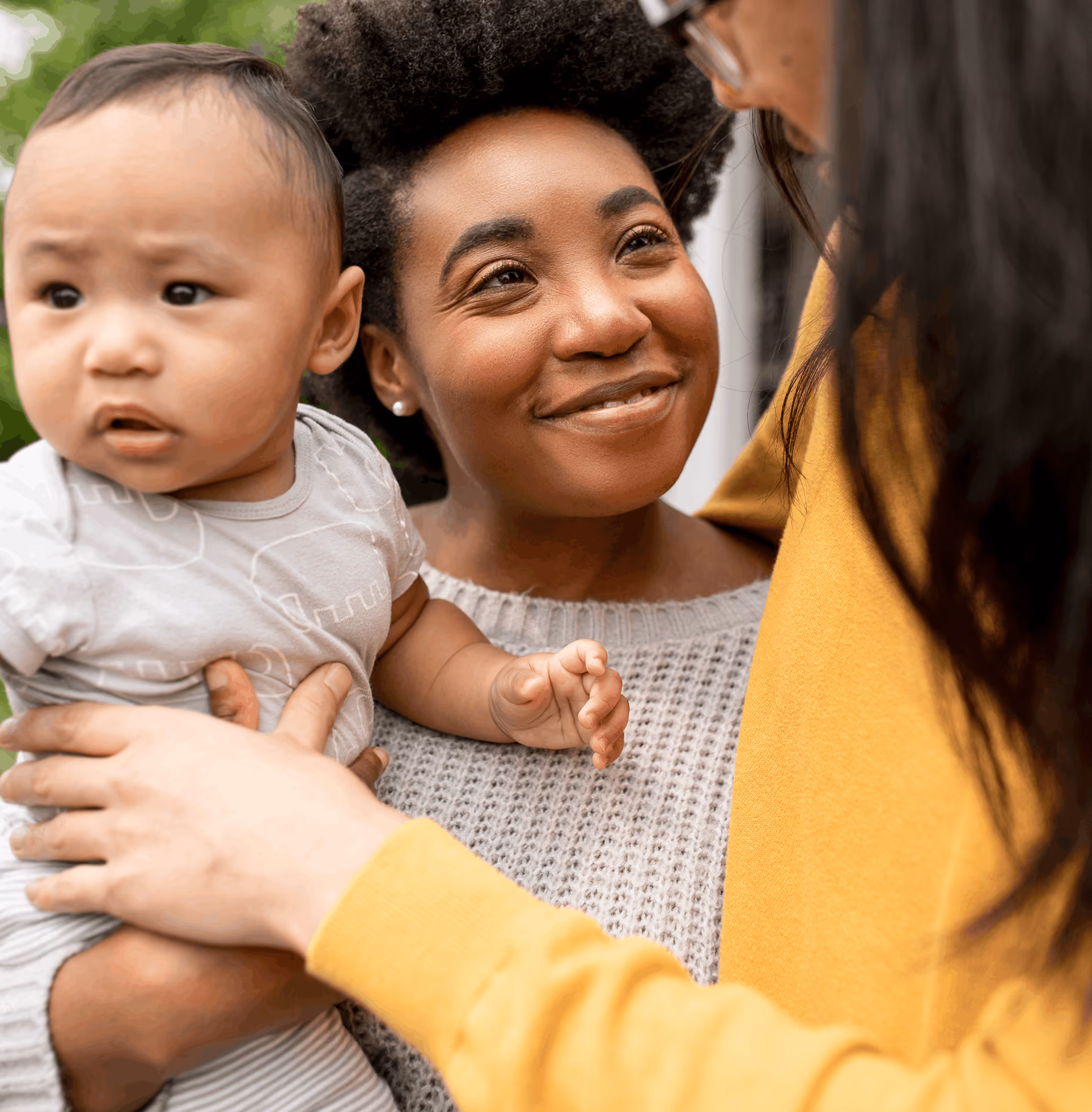 Smiling woman holding baby while another person in yellow reaches out to the baby outdoors.