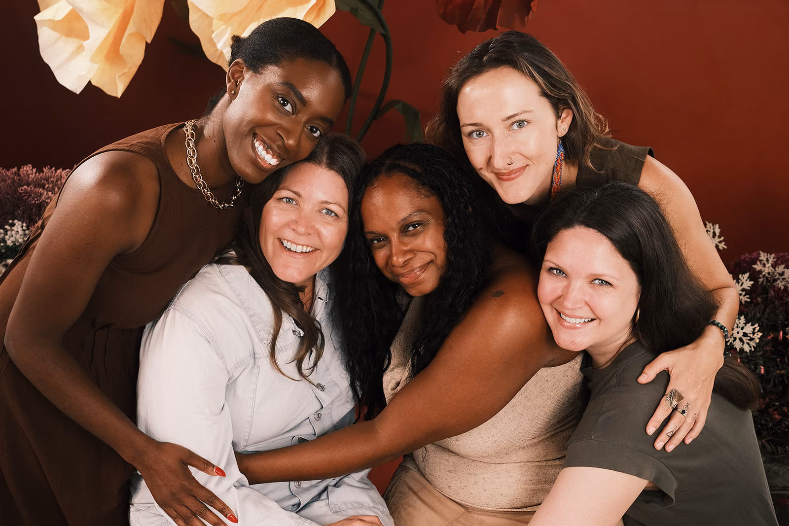 Three diverse women smiling and hugging each other outdoors in a sunny urban setting.