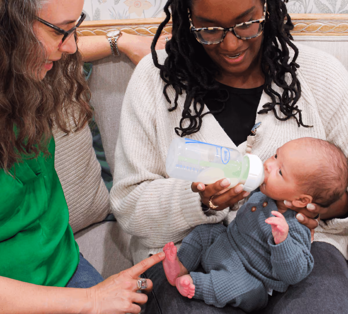 A woman wearing glasses and a white sweater feeds a baby in a blue outfit with a bottle while another woman in a green shirt smiles and gently touches the baby's foot on a gray couch.
