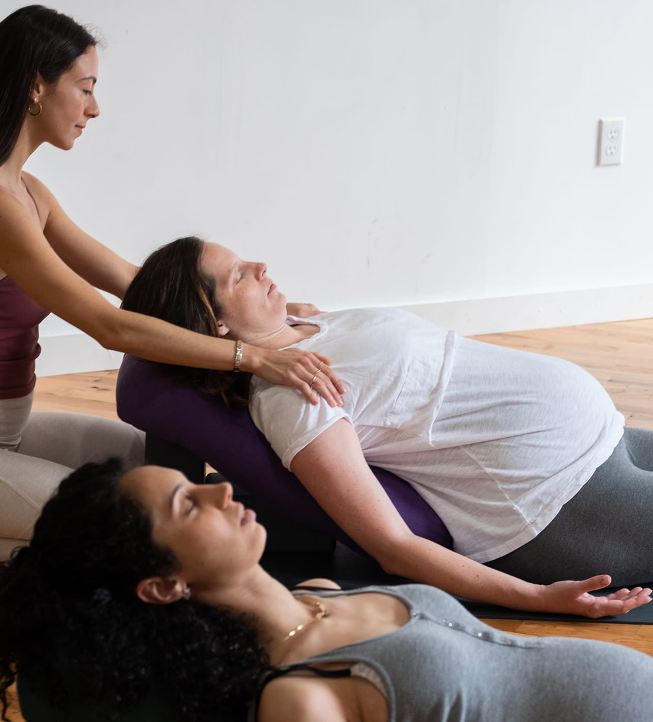 A pregnant woman lying back on a yoga bolster with eyes closed as an instructor gently supports her shoulders, while another person lies nearby, also relaxing with closed eyes in a yoga class.