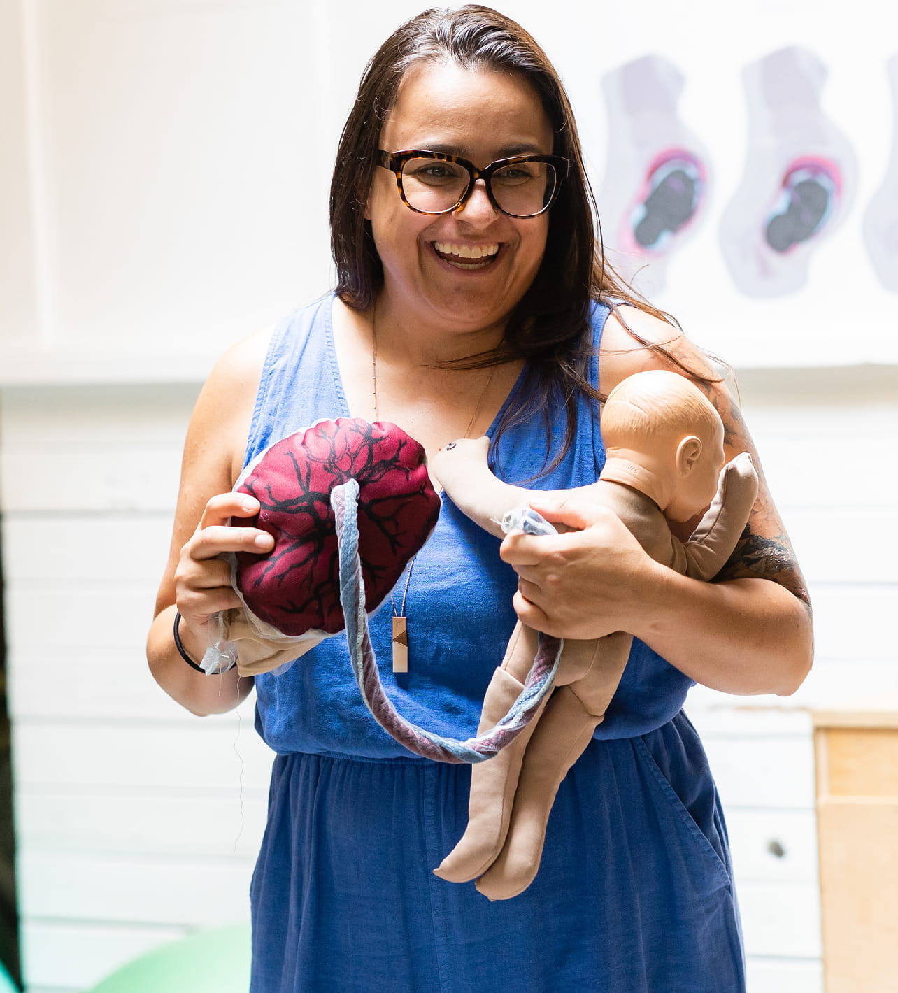 Smiling woman with glasses in a blue dress holding a baby doll connected by a fabric umbilical cord to a red fabric placenta model with black veins, demonstrating childbirth concepts.