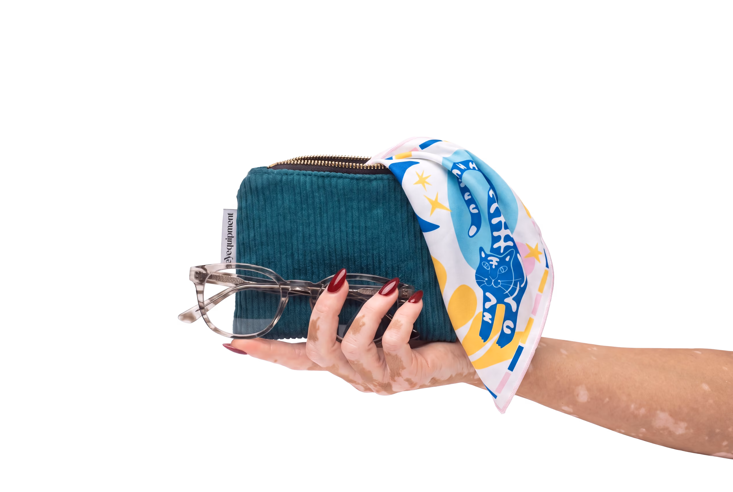 Hand mit roten Nägeln hält eine grüne Tasche, eine Brille und ein buntes Tuch mit Katzenmotiv.